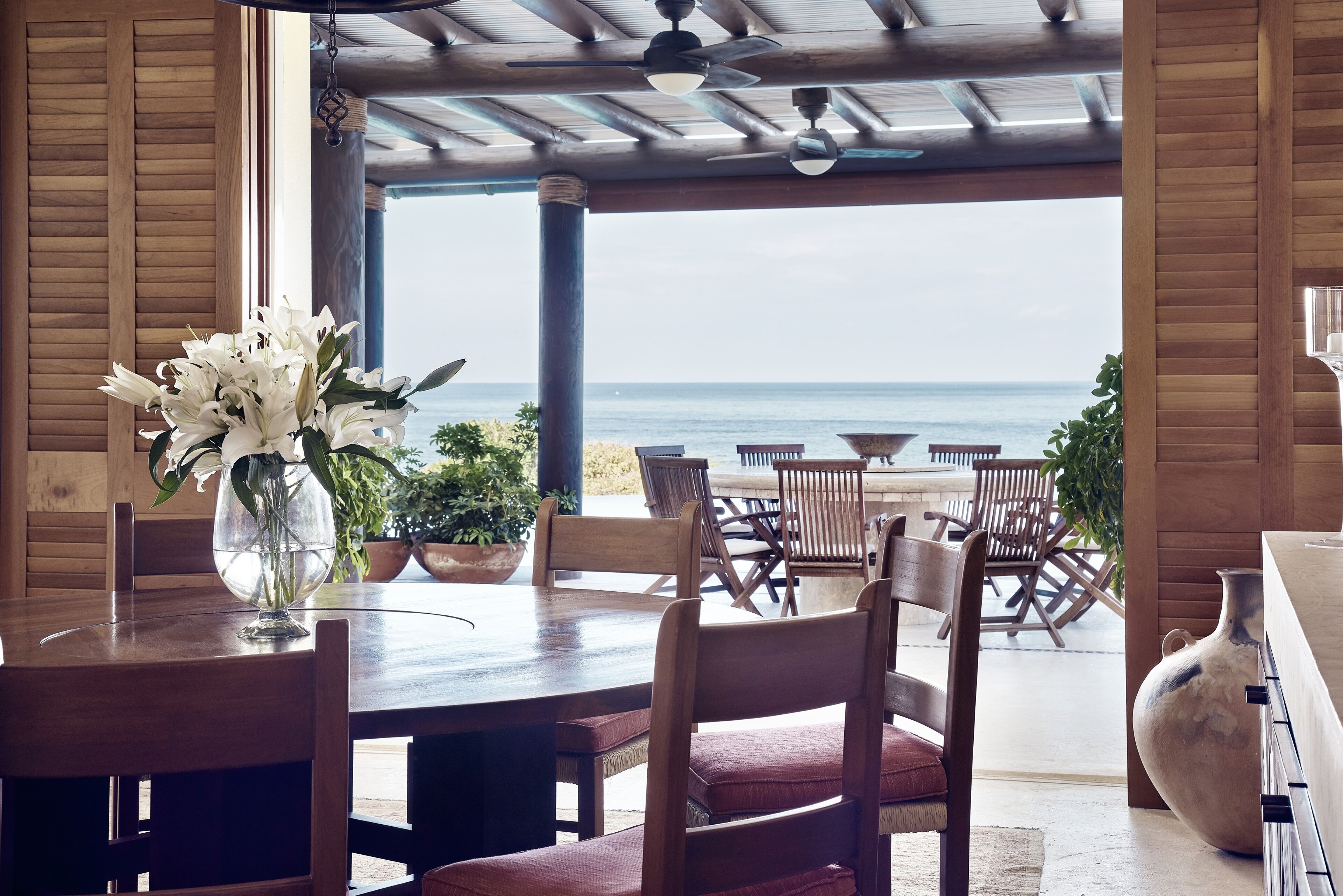 Un comedor de madera con vista al océano, un jarrón grande con flores y sillas de madera.