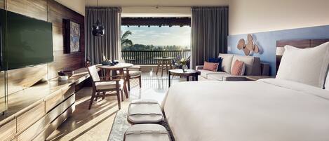 A hotel room with a large bed, a desk with a flat-screen TV, a balcony with a table and chairs, and a view of the ocean.