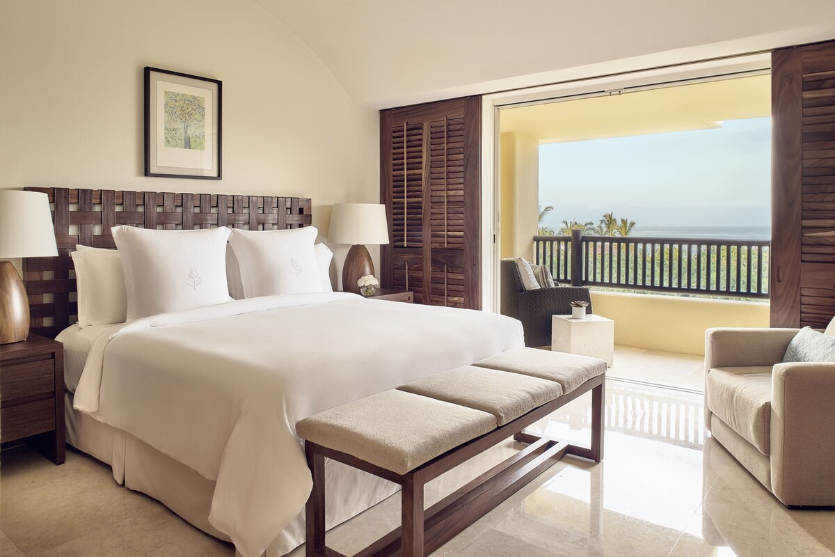 Ruime slaapkamer met een groot bed, nachtkastjes, een bankje en een balkon met uitzicht op zee.