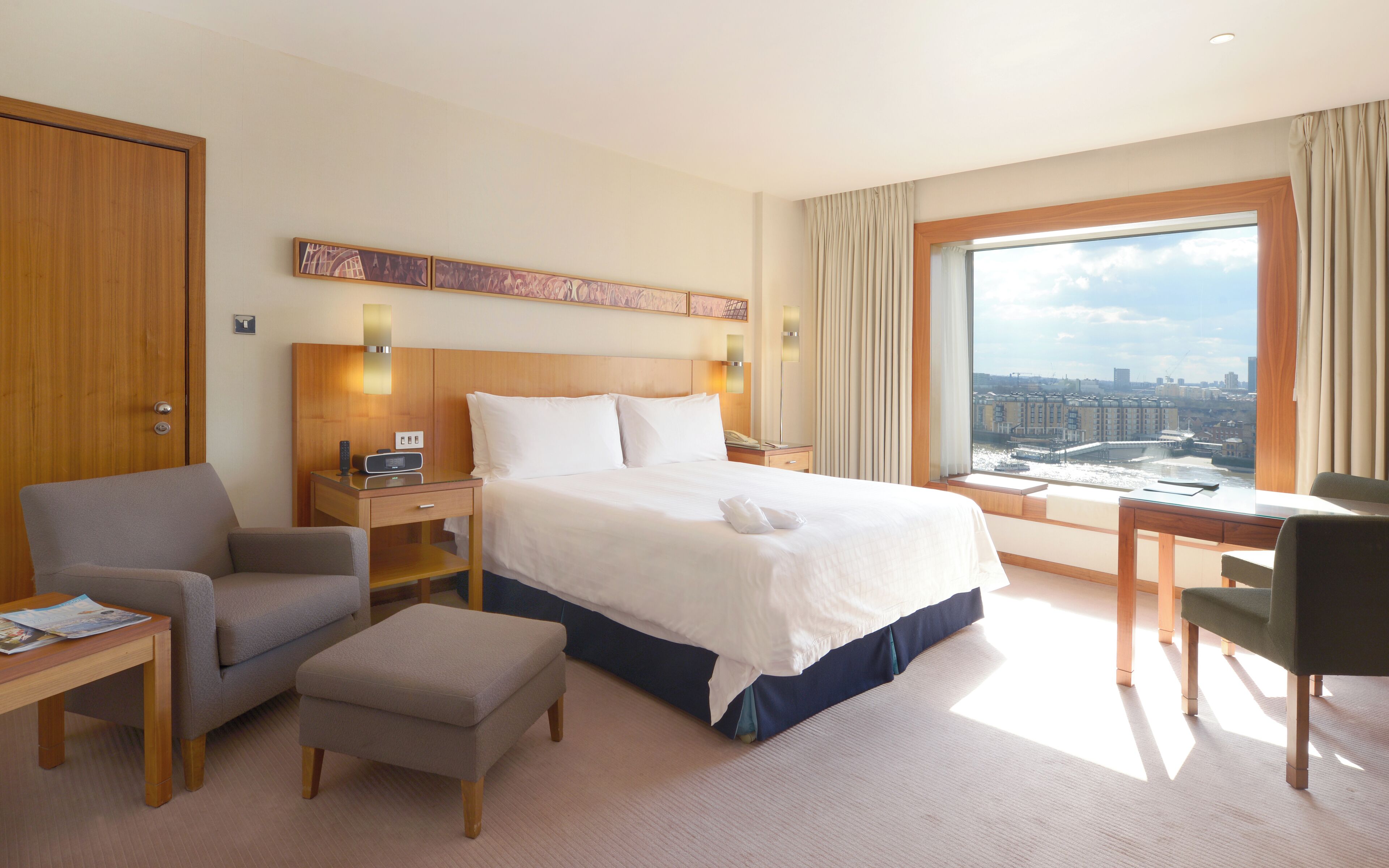 Een hotelkamer met een groot bed, een bureau, een stoel en uitzicht op een stadsgezicht door een groot raam.