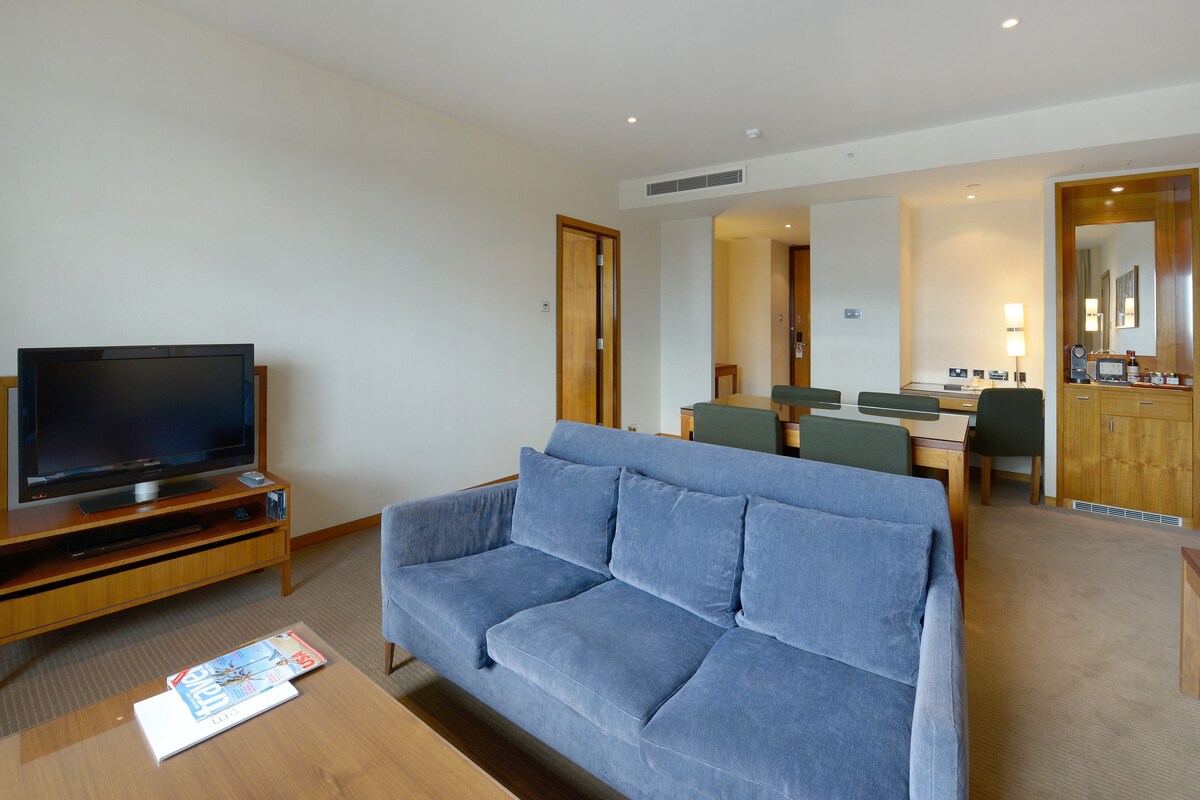 Une chambre d’hôtel moderne dotée d’une télévision à écran plat, d’un canapé bleu, d’un coin repas avec une table et des chaises, et d’un meuble en bois avec un miroir.