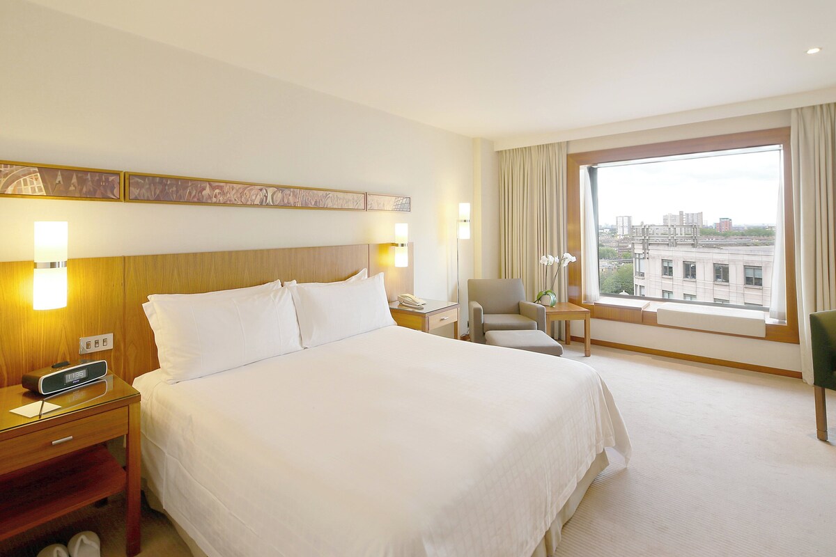 Une chambre d’hôtel avec un grand lit, des tables de chevet, une chaise, une petite table et une fenêtre donnant sur la ville.