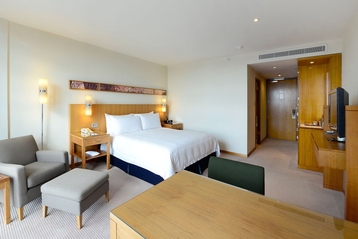 A hotel room with a bed, bedside table, lamp, chair, ottoman, and a TV.