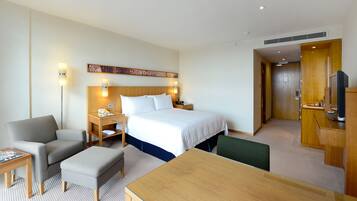 Una habitación de hotel con cama, mesita de noche, lámpara, sillón, reposapiés y televisor.