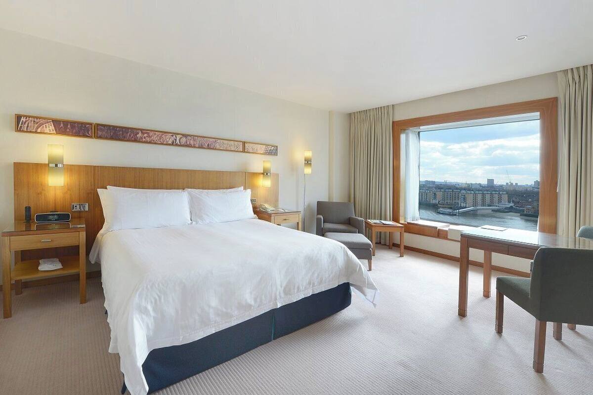 A hotel room with a large bed, a desk, a chair, and a view of a cityscape through a large window.