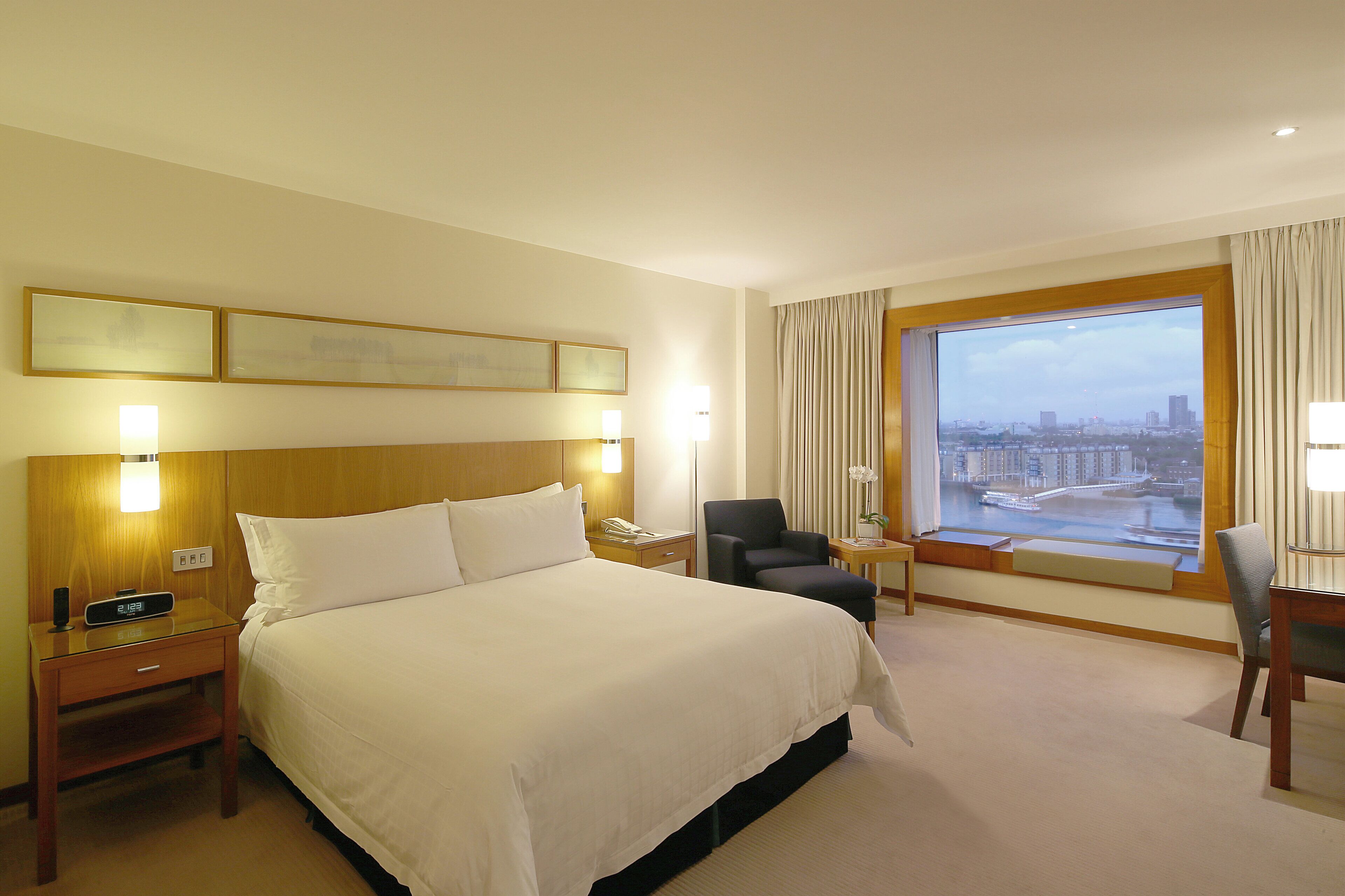 A hotel room with a large bed, a desk with a chair, a nightstand, a window with a city view, and a wall-mounted lamp.