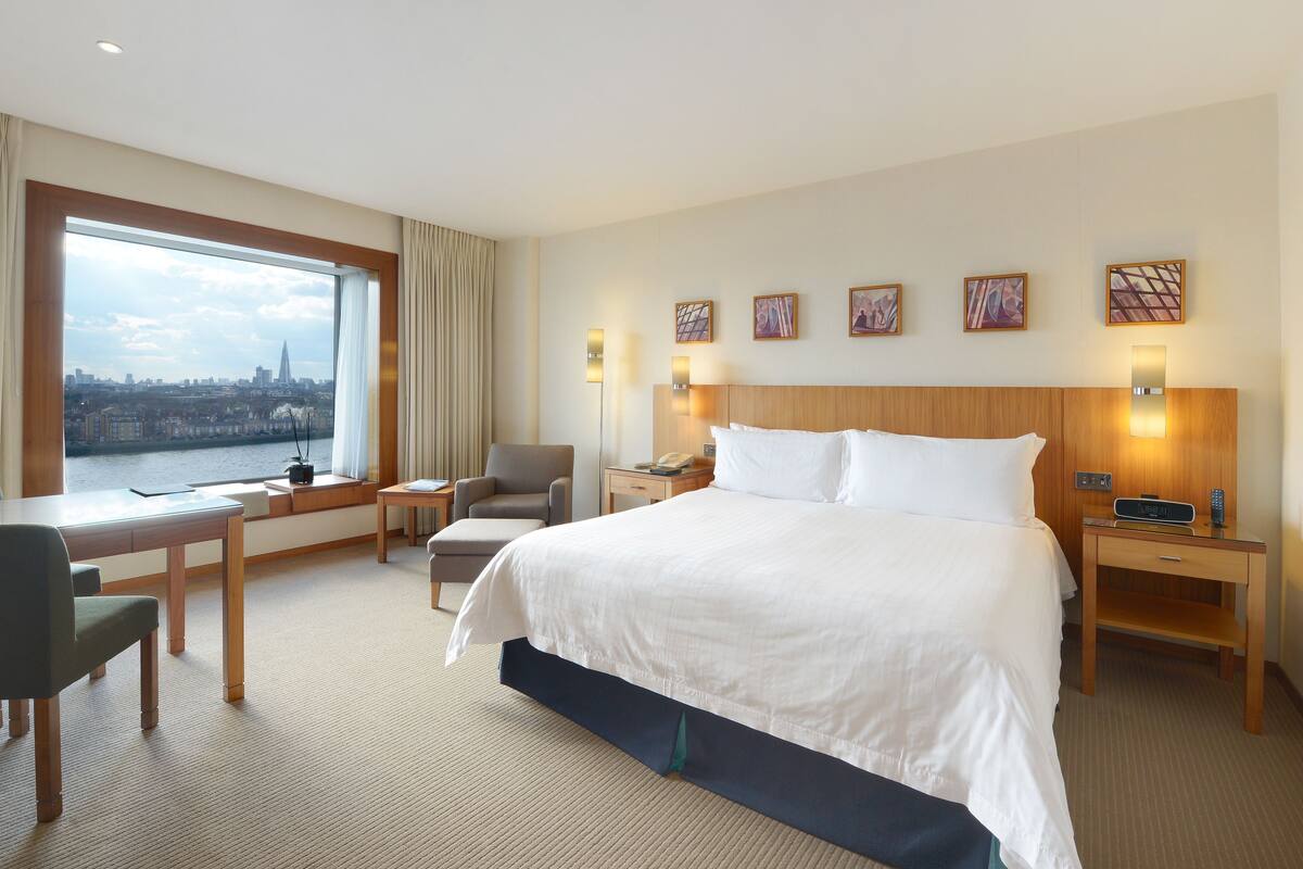 A hotel room with a large bed, a desk, a chair, a nightstand, and a view of a cityscape through a large window.