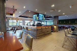 Zona de bar moderna con mostrador de madera, diversas opciones de asientos, incluyendo taburetes y mesas, y grandes televisores de pantalla plana montados en el techo.