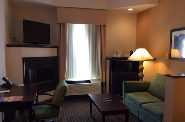 A hotel room with a flat-screen TV mounted on the wall, a sofa, a small table, a lamp, and a window with curtains. - Wingate by Wyndham High Point/Greensboro/Airport South (High Point)