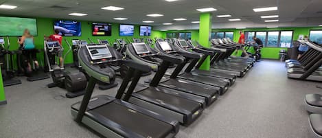 A gym with multiple treadmills, exercise bikes, and a green wall with screens.