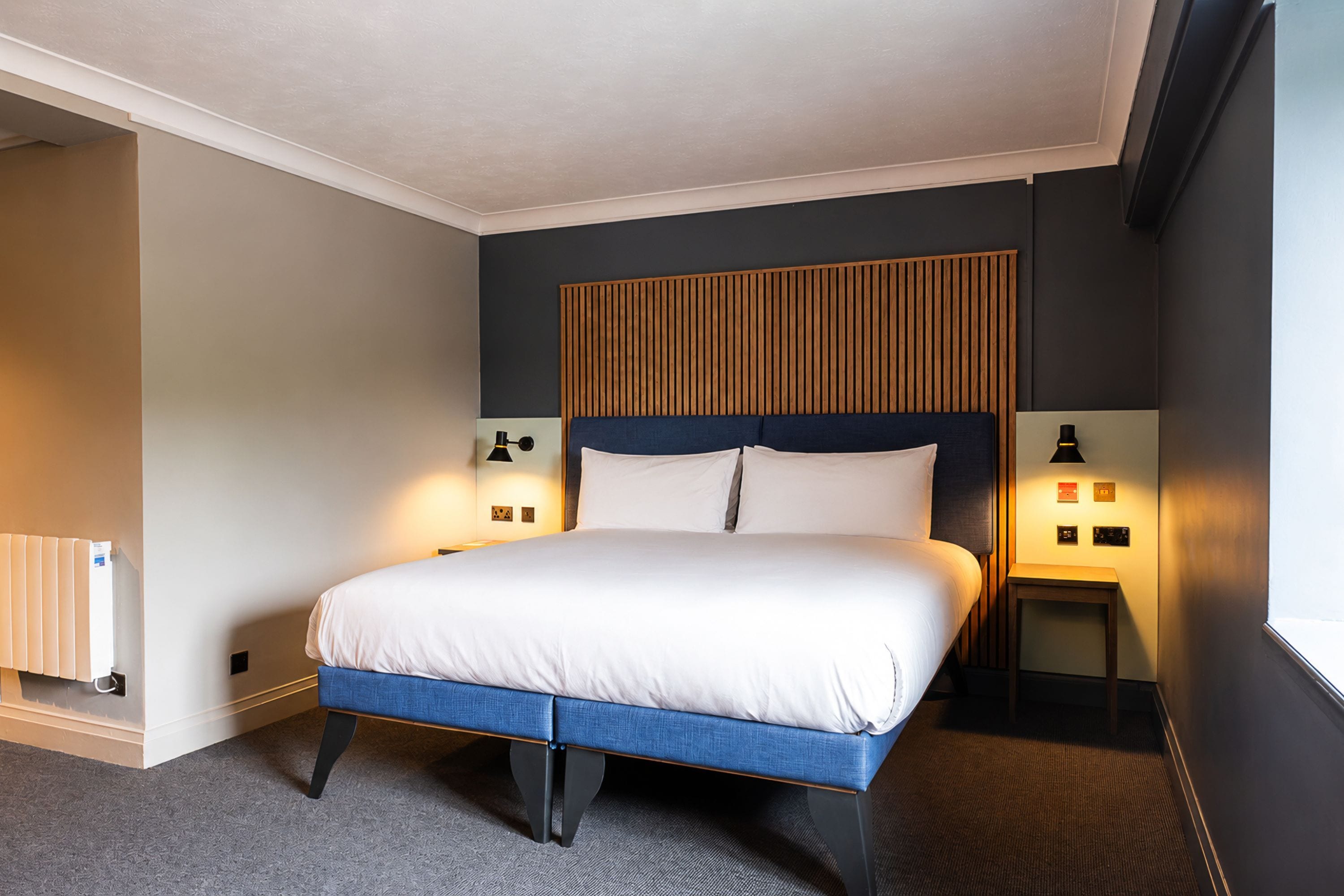 A hotel room with a bed, bedside lamps, a radiator, and a window.
