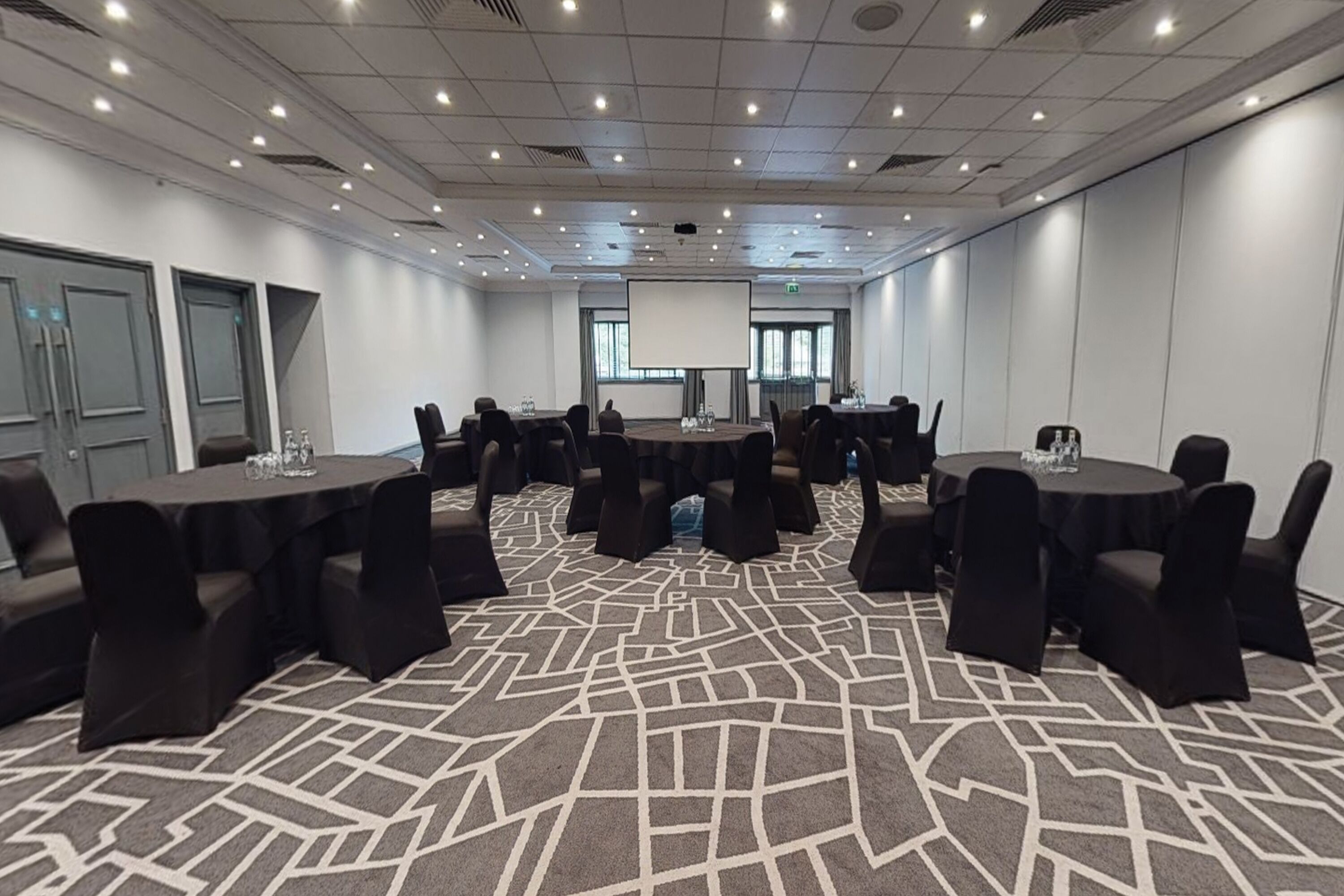 A conference room with multiple tables and chairs, a projector screen, and a patterned carpet.