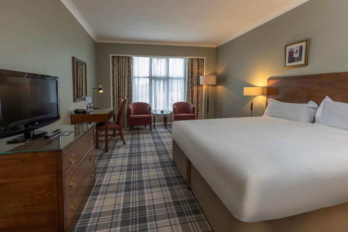 A hotel room with a bed, a television, a desk with a chair, and a window with curtains.