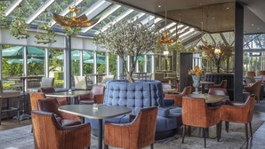Un espace intérieur moderne avec un grand canapé bleu, des chaises et des tables en bois. De grandes fenêtres illuminent la pièce, et un arbre se dresse au centre.