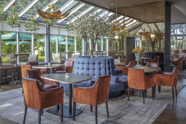 A modern indoor space with a large blue sofa, wooden chairs, and tables. There are large windows and a tree in the center.