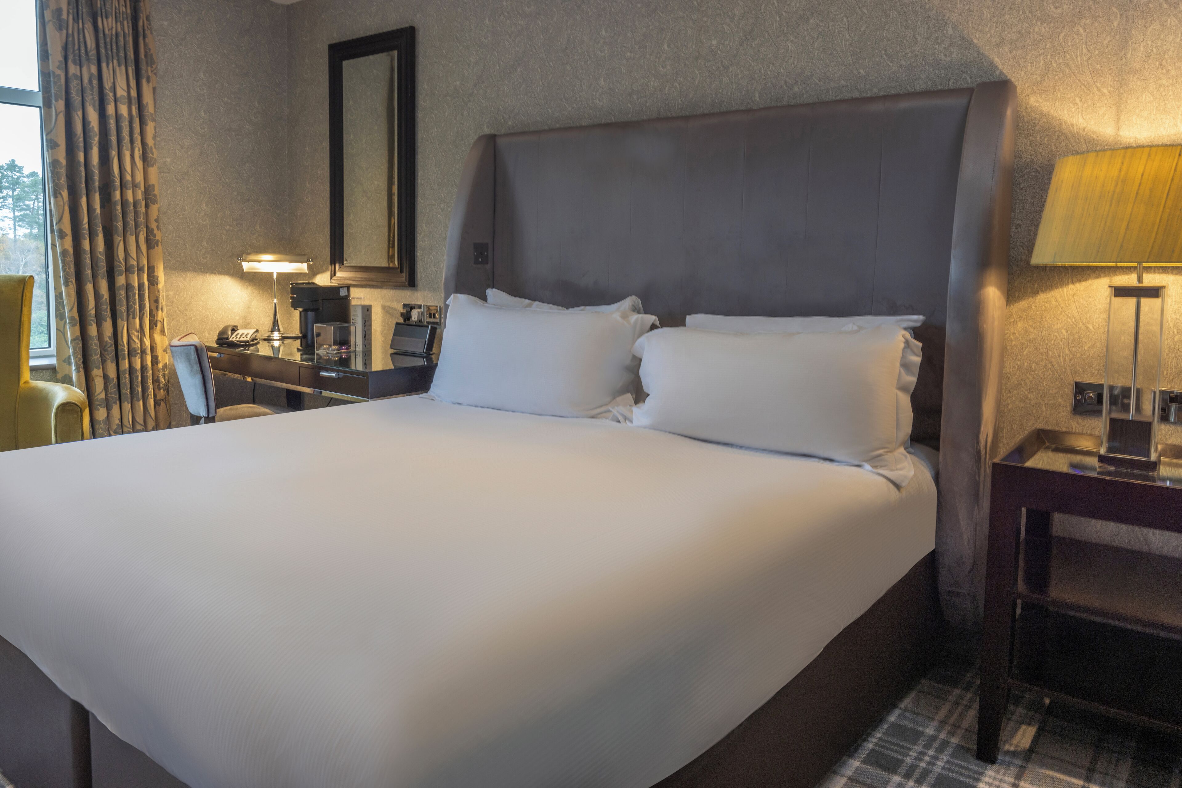 Een hotelkamer met een groot bed, een bureau met een lamp, een stoel en een raam met gordijnen.