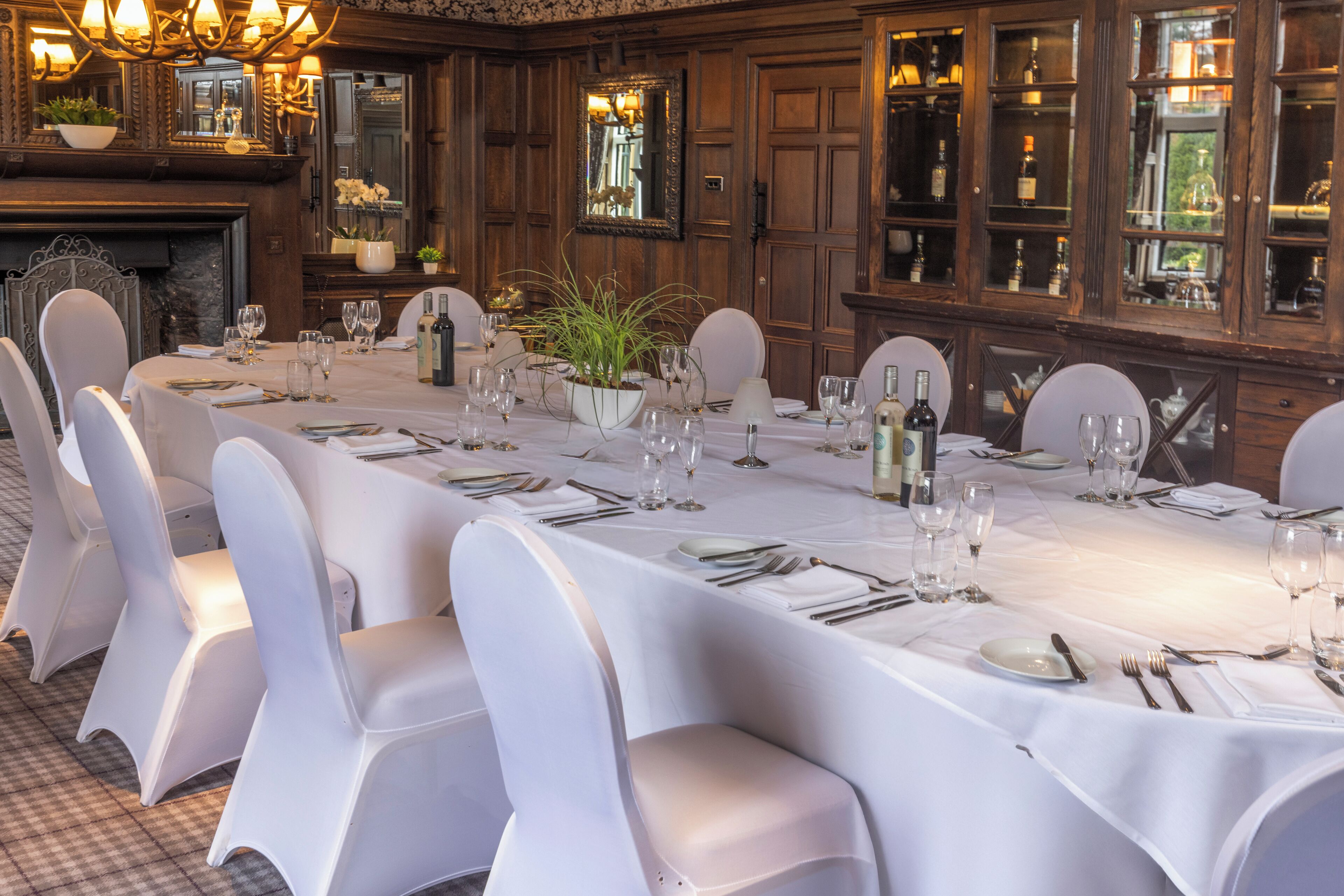 Een lange eettafel gedekt voor een formeel diner, met witte stoelen, een kroonluchter en houten lambrisering.