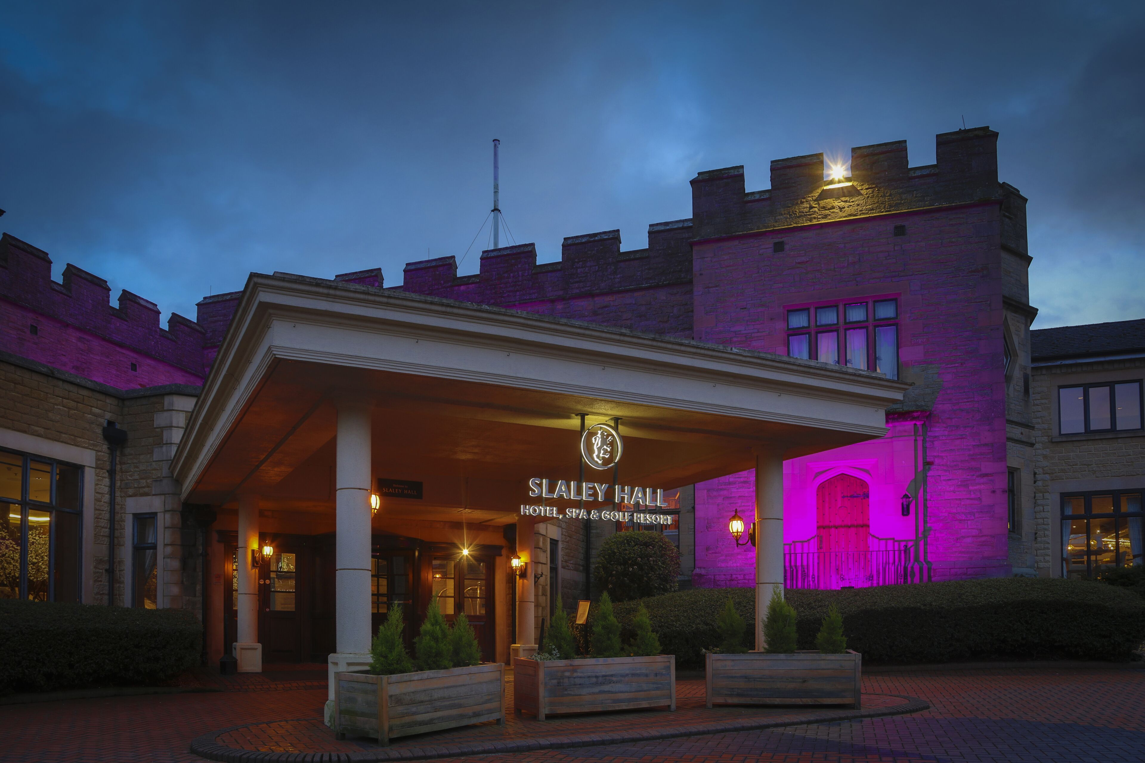 L’entrée de l’hôtel Slaley Hall, avec sa façade en briques, son espace d’entrée abrité et un panneau indiquant qu’il s’agit d’un hôtel, d’un centre de bien-être et d’un complexe de golf.