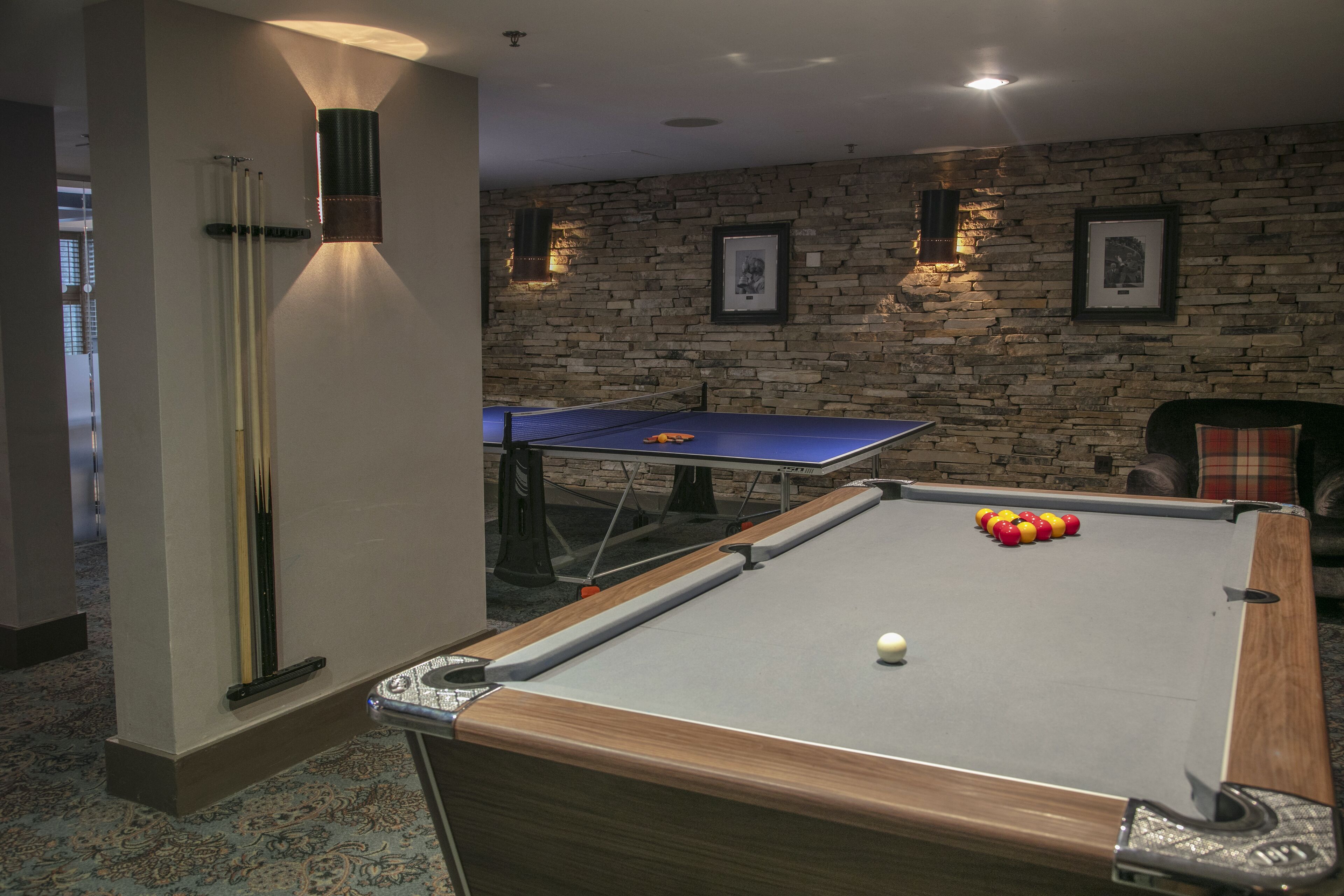 A pool table with a cue stick, a billiards table, and a billiard ball in a room with stone walls and framed pictures.