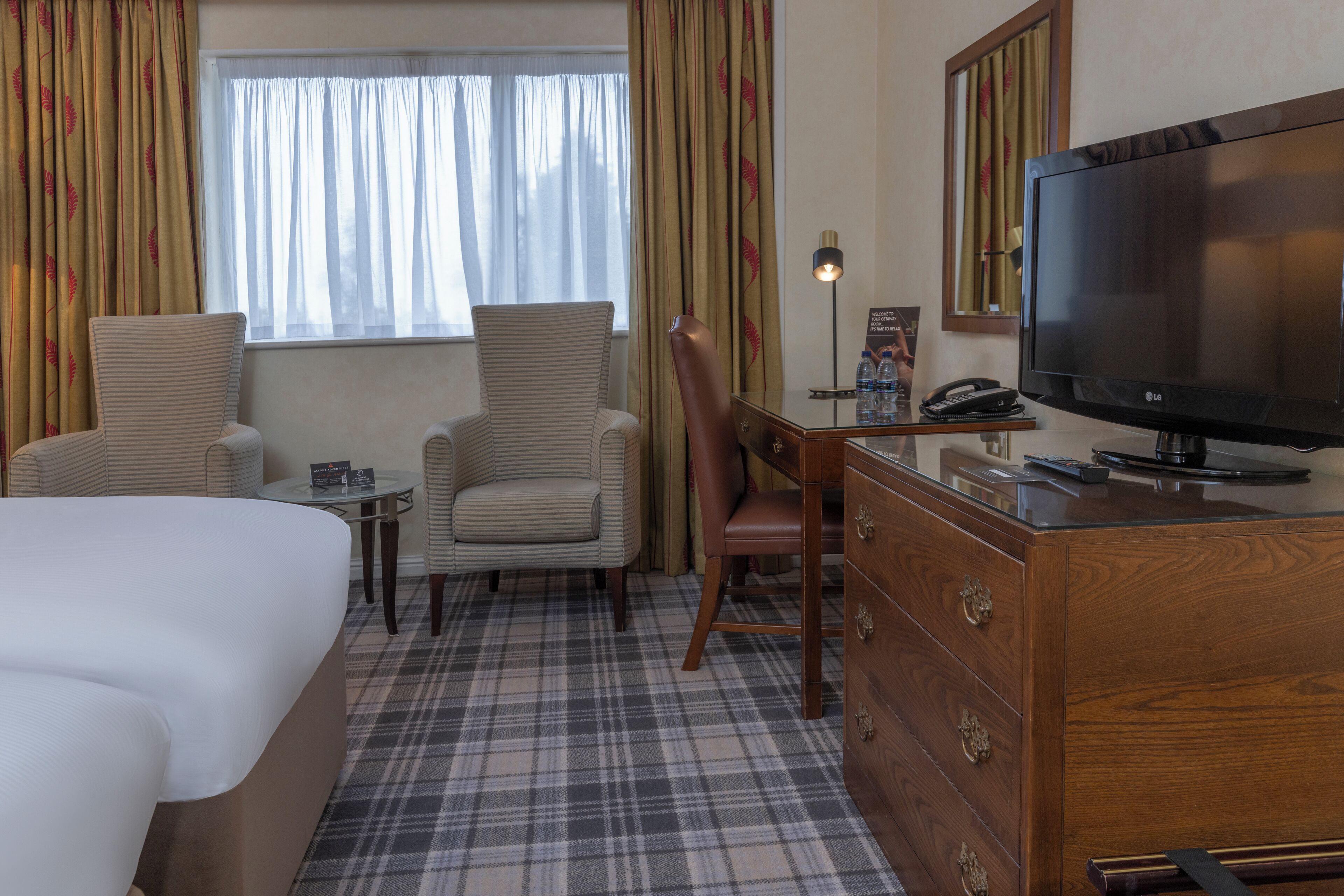 Ett hotellrum med en säng, två stolar, ett skrivbord med en tv, en byrå och ett fönster med gardiner.
