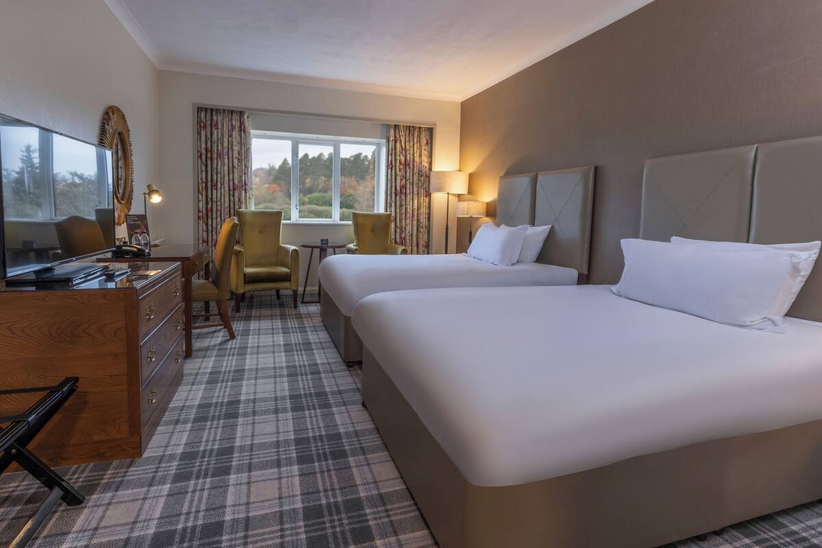 A hotel room with two beds, a desk with a computer, a chair, a small table, and a window with curtains.