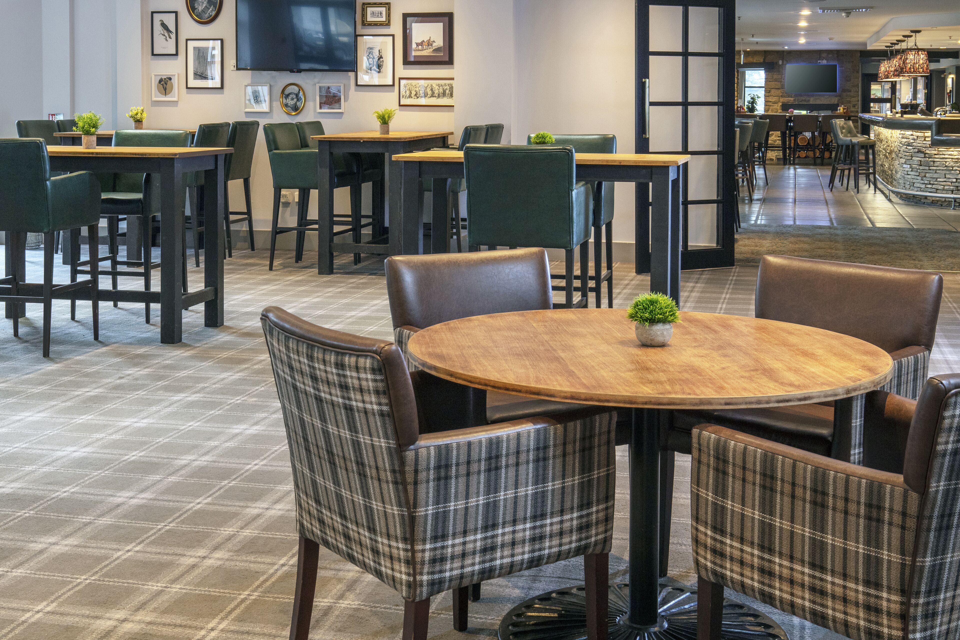 A modern dining area with round tables and plaid chairs, a flat-screen TV on the wall, and a bar area in the background.