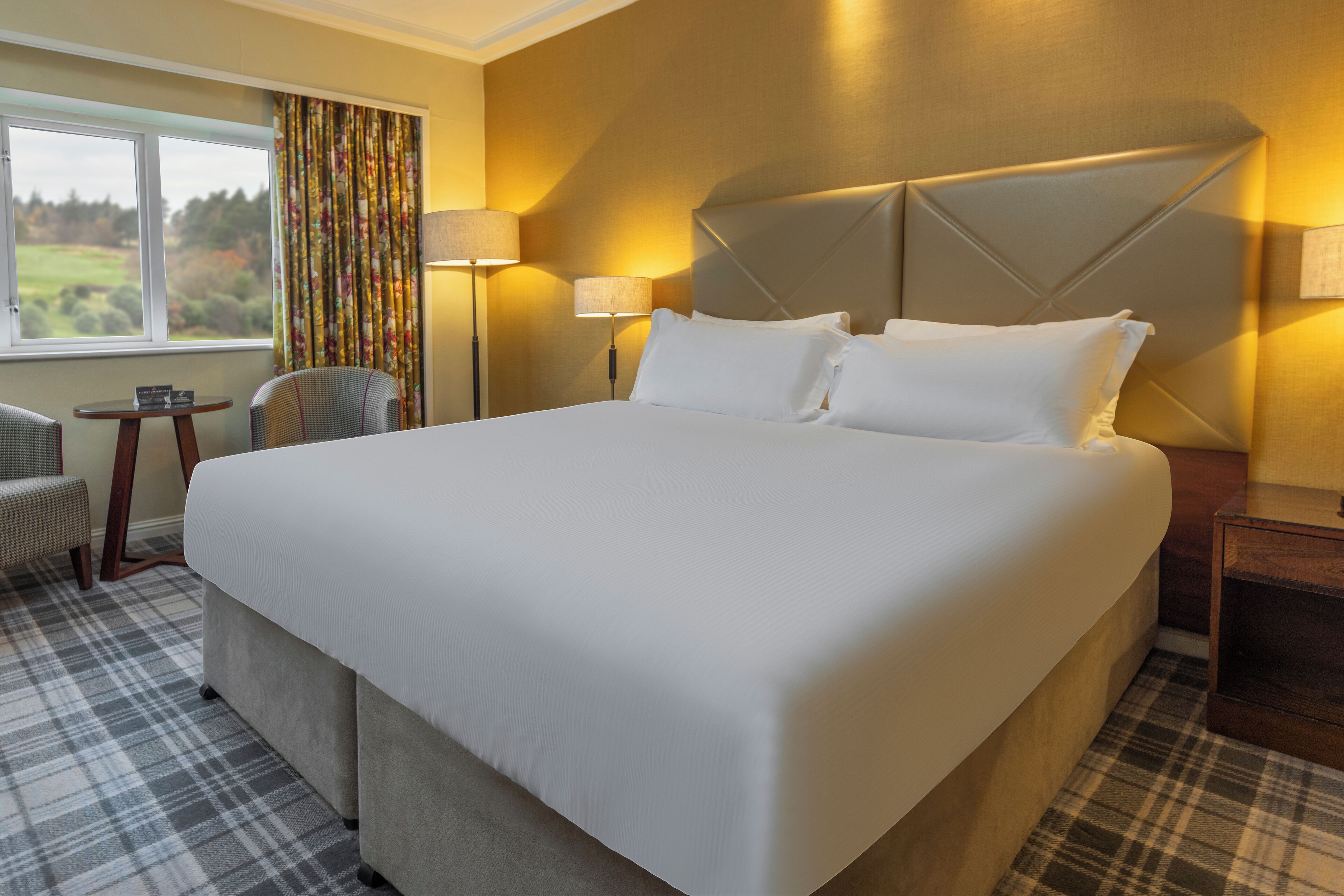 A hotel room with a large bed, two bedside lamps, a chair, a small table, and a view of the outdoors through a window.