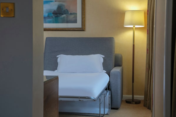 Une chambre d’hôtel avec un lit, un fauteuil gris, un lampadaire et un tableau encadré au mur.