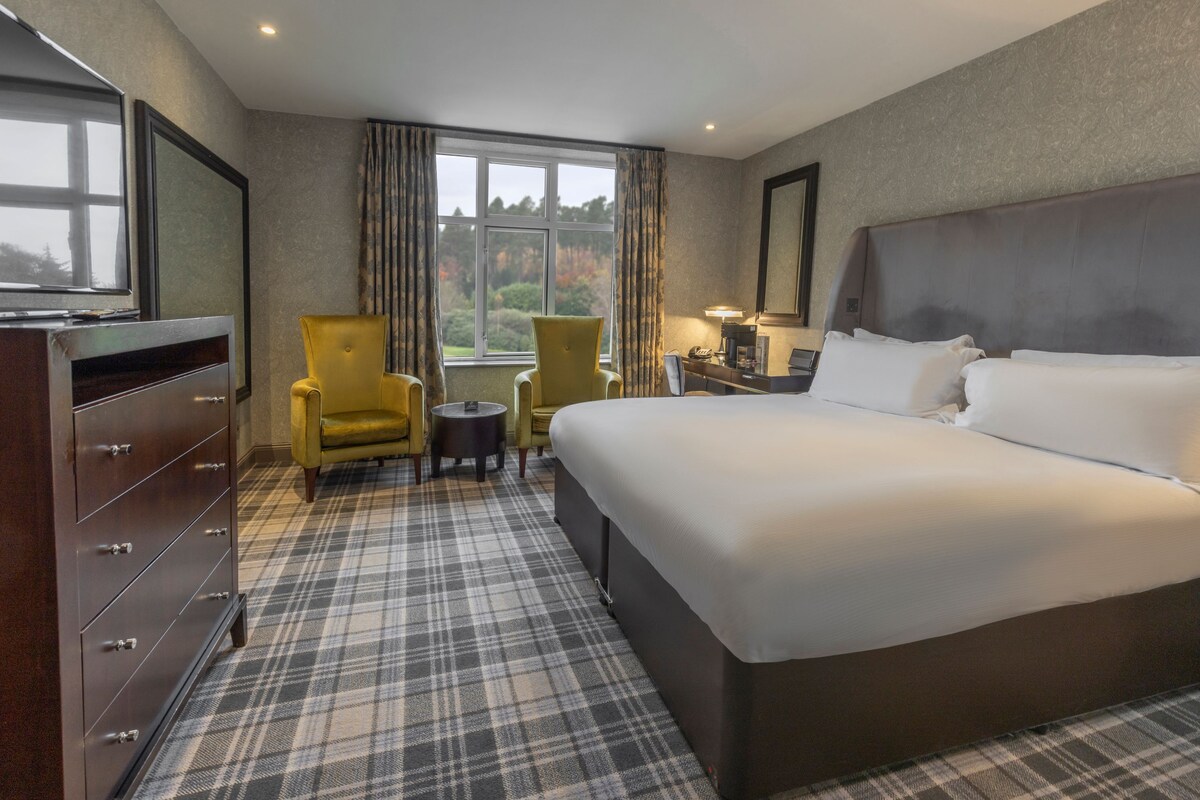 A hotel room with a large bed, a dresser, a chair, a small table, and a window with a view.