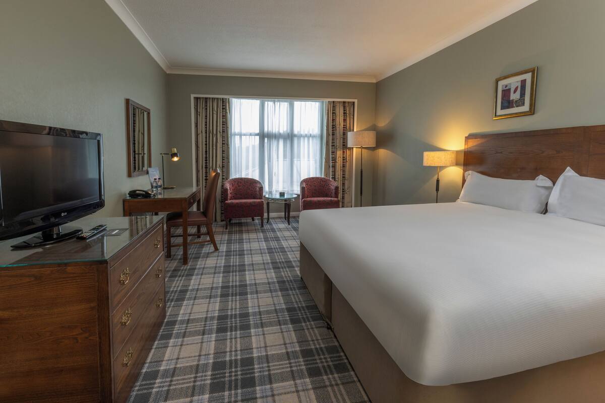 A hotel room with a large bed, a desk with a television, two chairs, a small table, and a window with curtains.