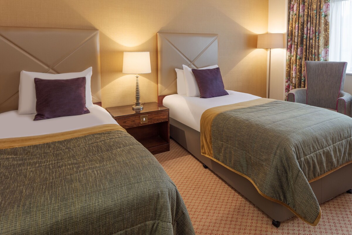 A hotel room with two beds, a nightstand, a chair, and a window with curtains.