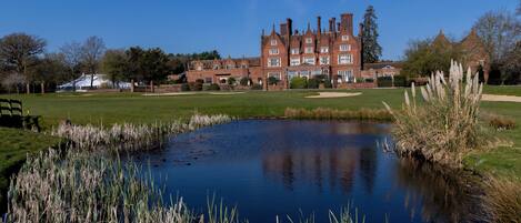 A large brick building with multiple chimneys, a pond with tall grasses, and a golf course in the foreground.