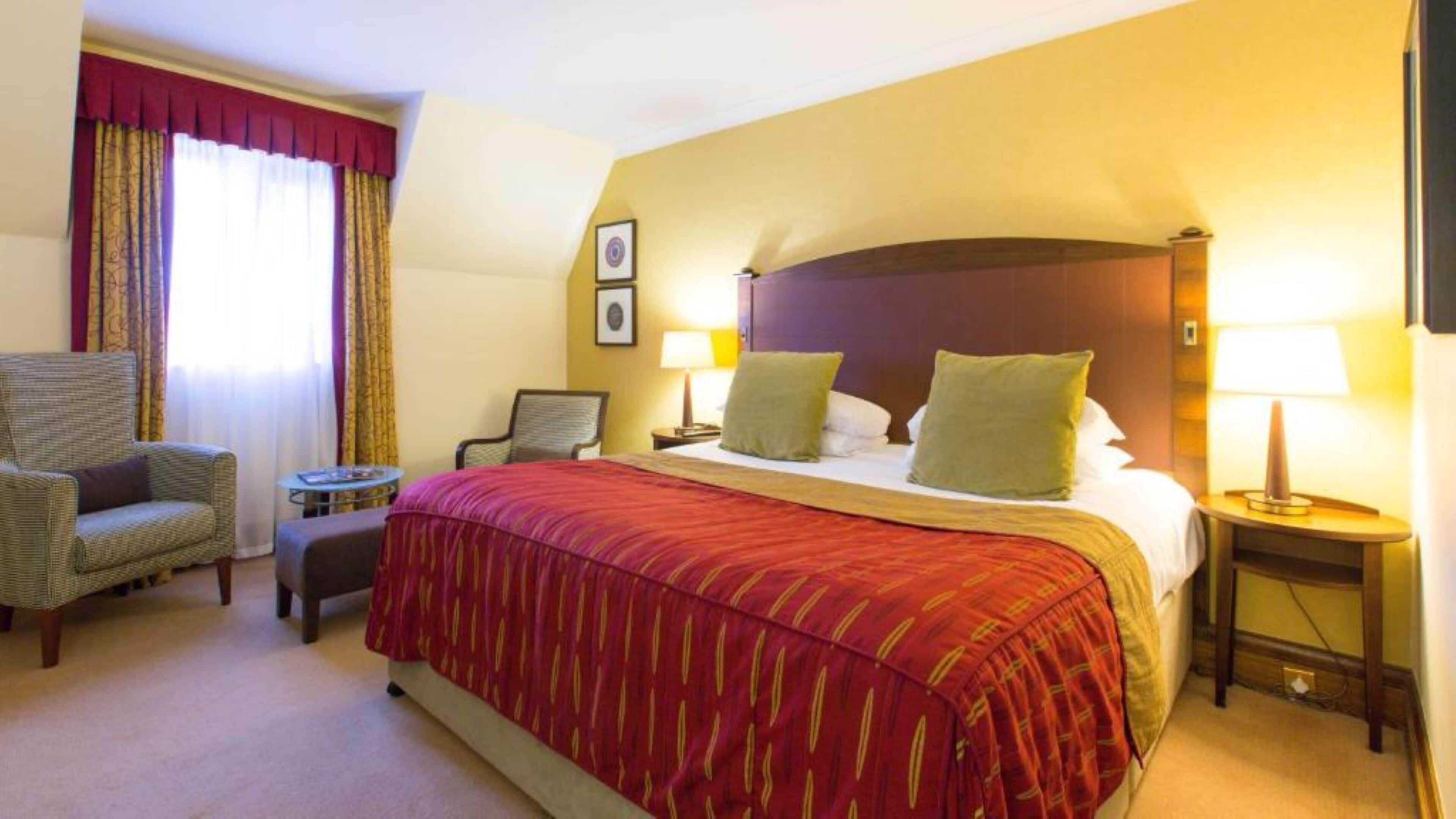 Ein Hotelzimmer mit einem großen Bett, zwei Nachttischlampen, einem Sessel, einem kleinen Tisch und einem Fenster mit Vorhängen.