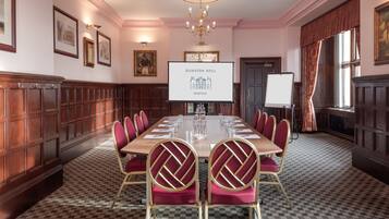 A conference room with a long table, red upholstered chairs, a chandelier, and framed pictures on the walls.