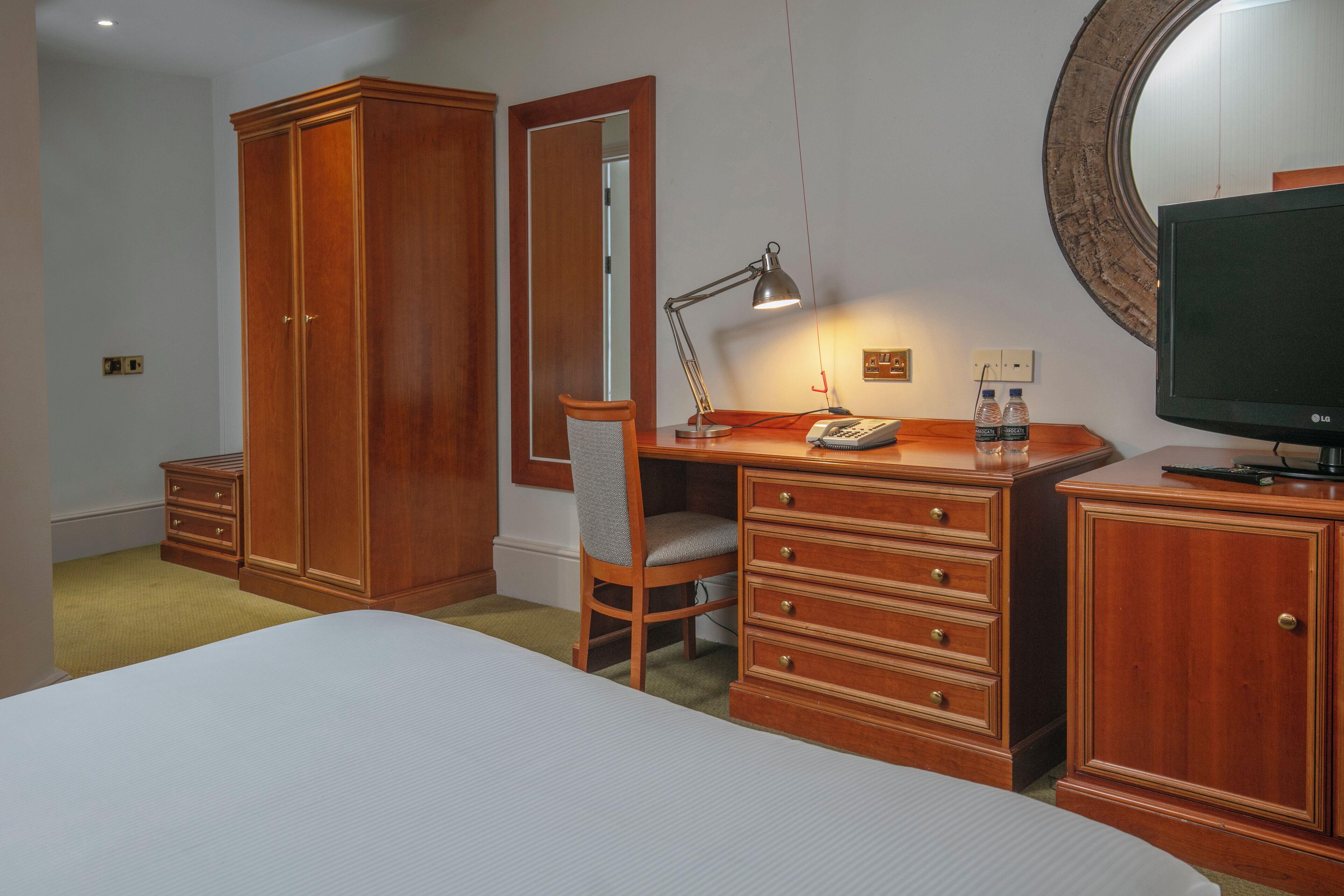 A bedroom with a bed, wooden wardrobe, desk with chair, desk lamp, mirror, and a television.