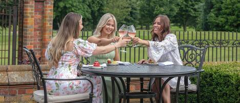 Three people toasting at a round metal table outdoors with a brick structure and a metal fence in the background.