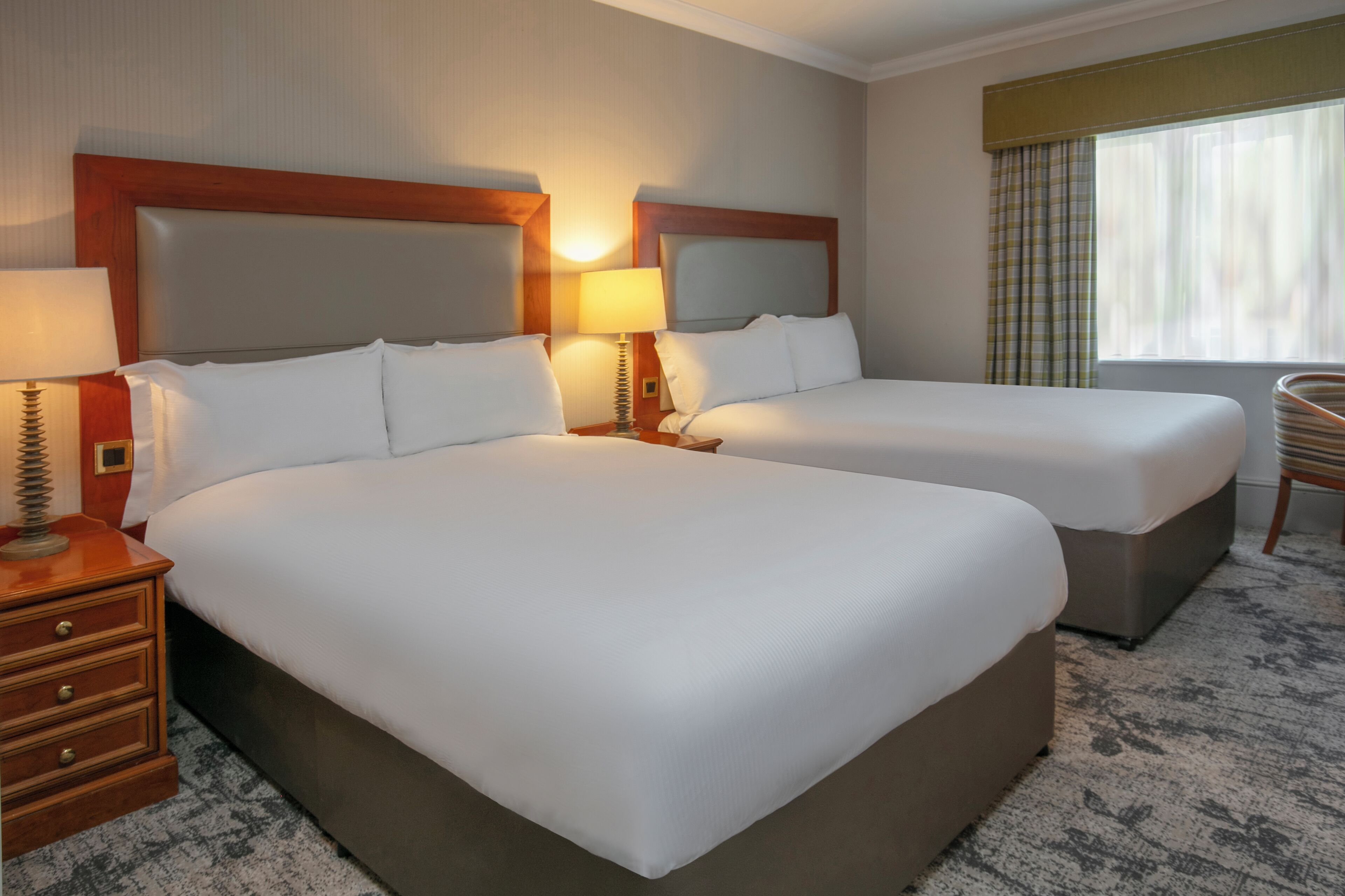 A hotel room with two beds, a wooden headboard, a bedside table with a lamp, and a window with curtains.
