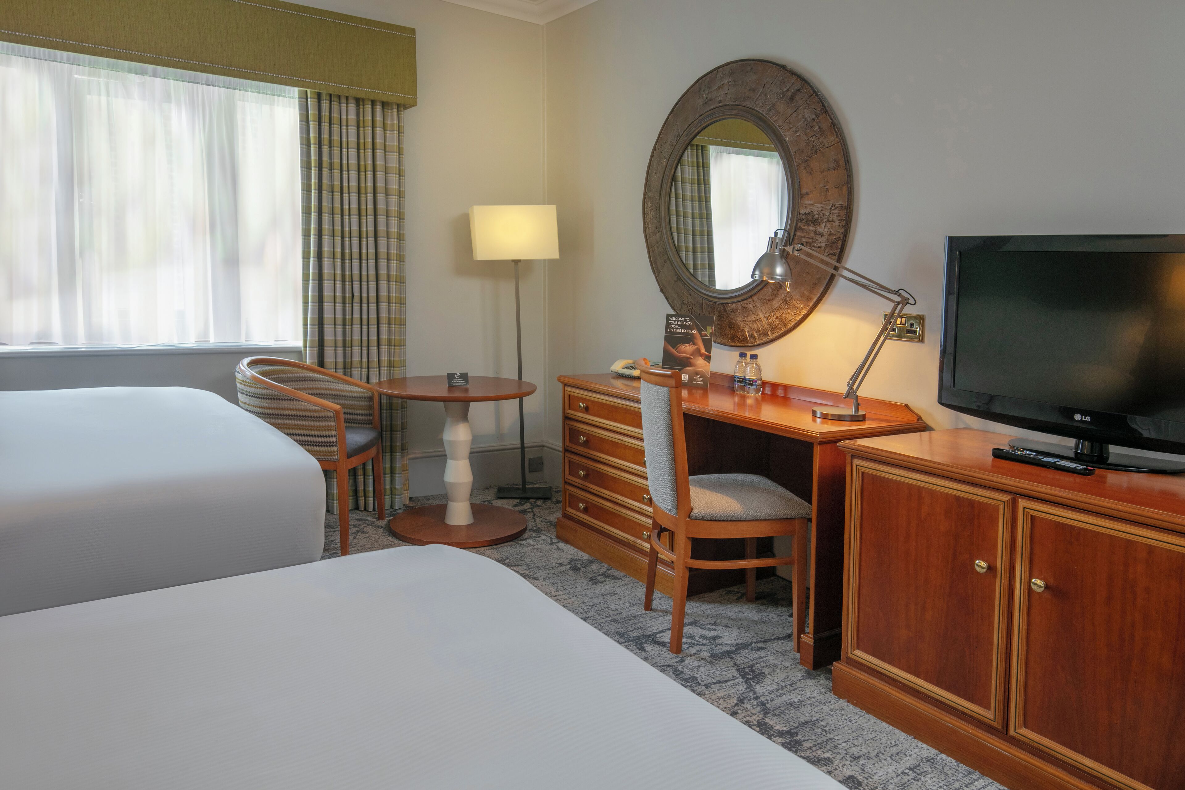 A hotel room with two beds, a desk with a chair, a television, a lamp, and a large mirror.