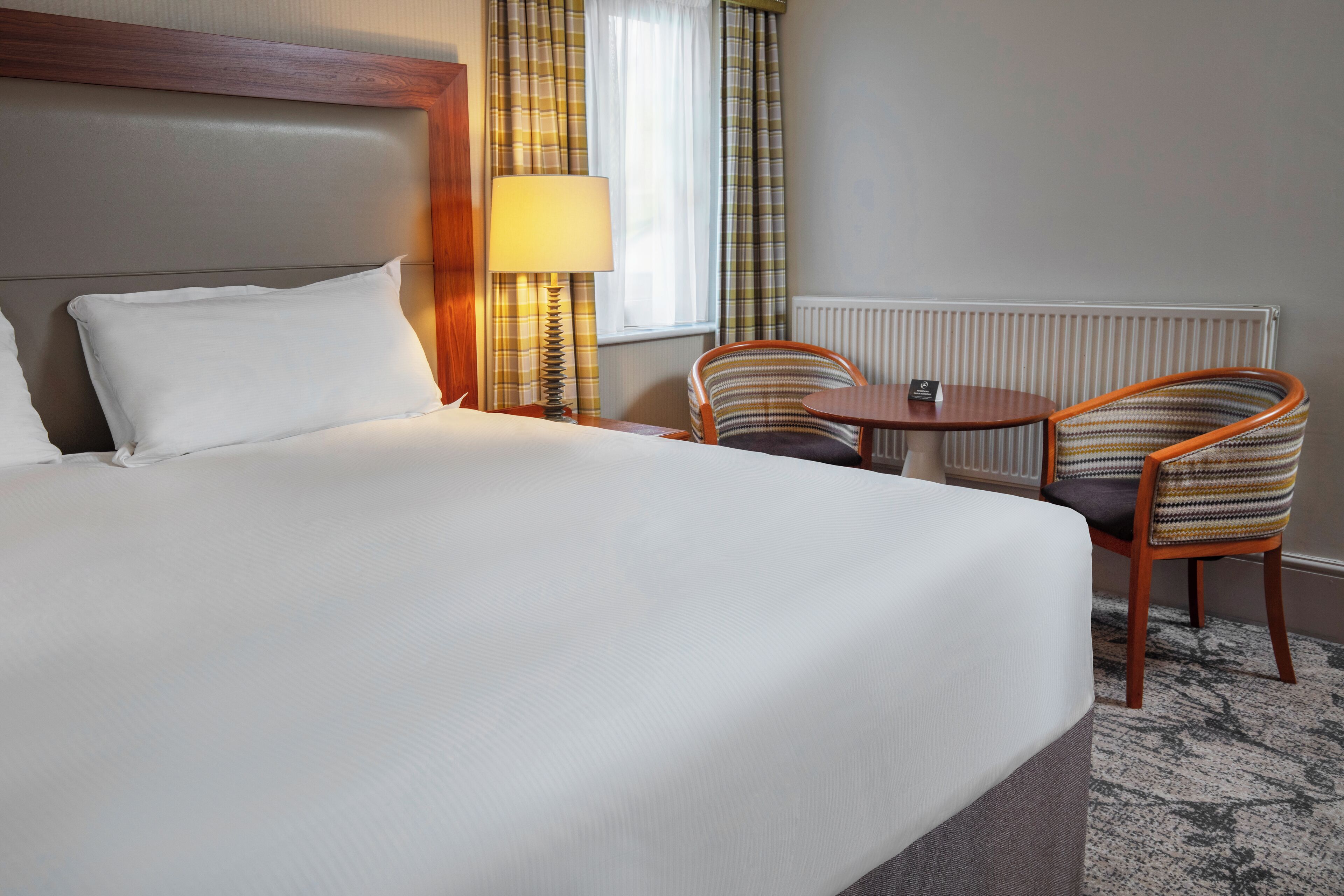 A hotel room with a large bed, two chairs, a small table, a lamp, and a window with curtains.