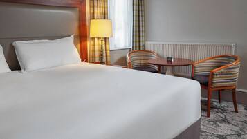 A hotel room with a large bed, two chairs, a small table, a lamp, and a window with curtains.