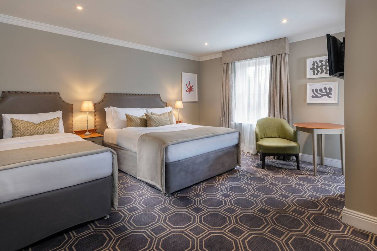 A hotel room with two beds, a green armchair, a small table, and artwork on the walls.
