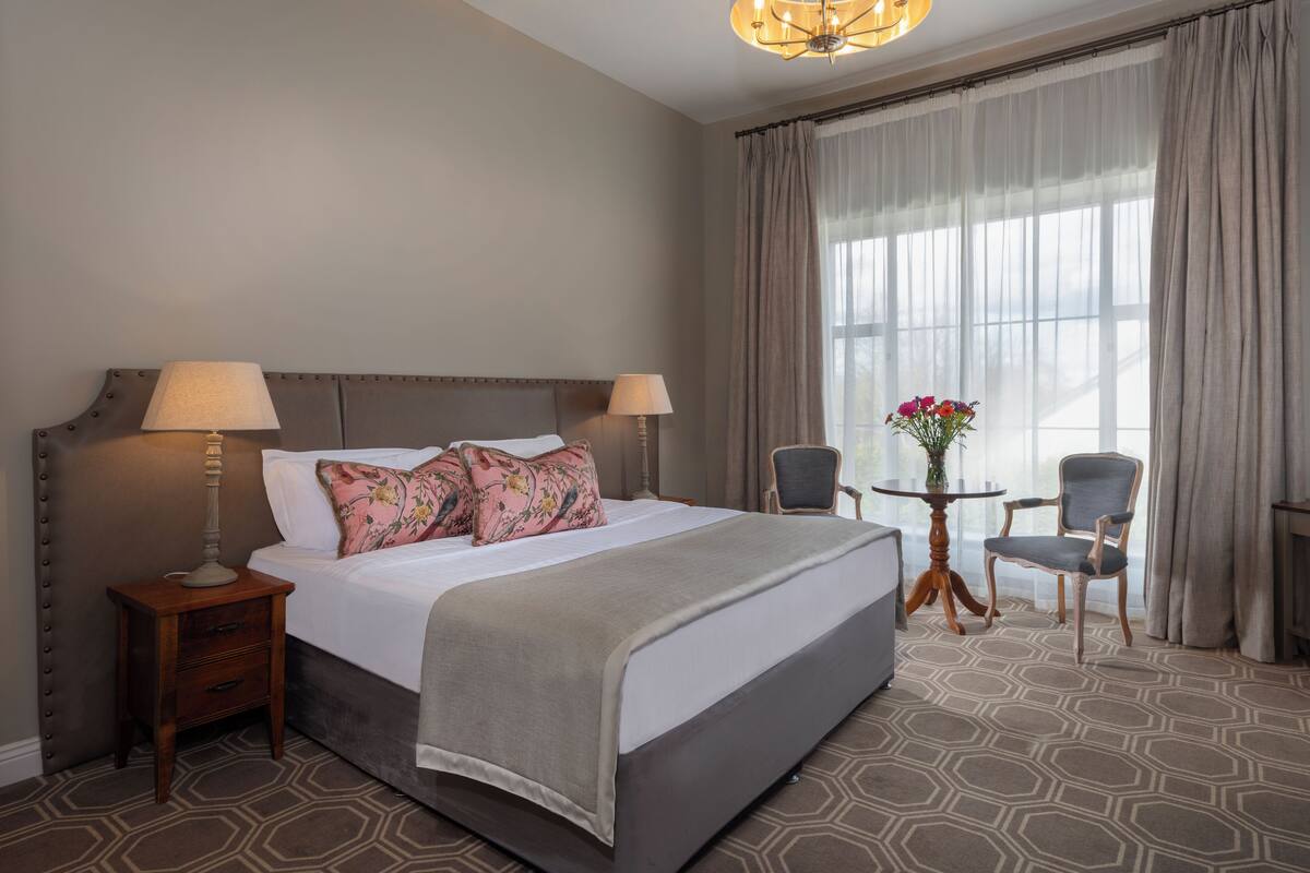 A hotel room with a large bed, two bedside lamps, a small table with a vase of flowers, two chairs, and a large window with curtains.