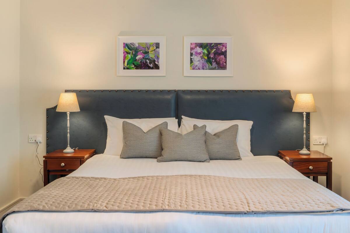 A neatly made bed with a quilted cover, two bedside tables with lamps, and two framed floral paintings on the wall.