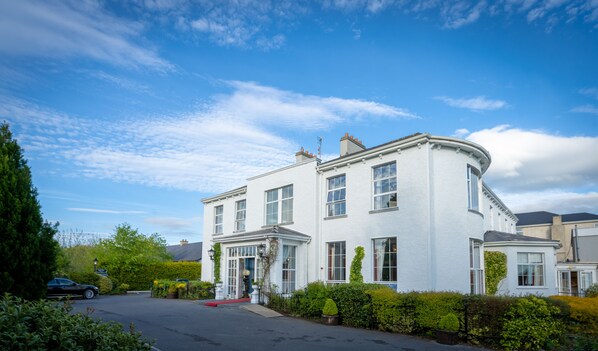 A white two-story house with a curved corner, multiple windows, and a red carpet leading to the entrance. - Castle Oaks House Hotel (Castleconnell)