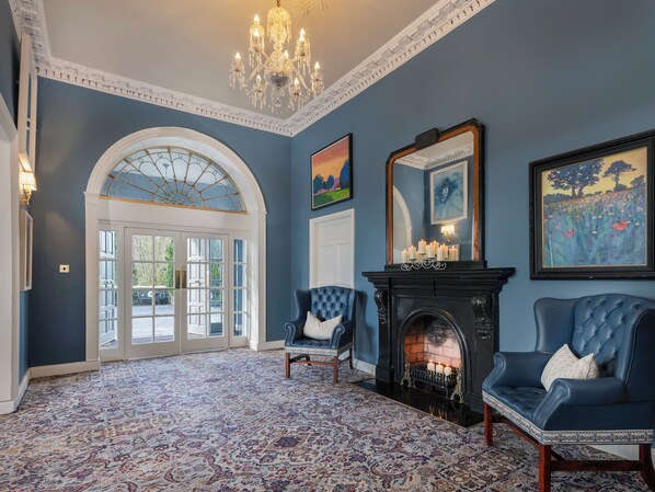 A room with a patterned carpet, a fireplace with candles, two blue upholstered chairs, a chandelier, and a large arched doorway. - Castle Oaks House Hotel (Castleconnell)