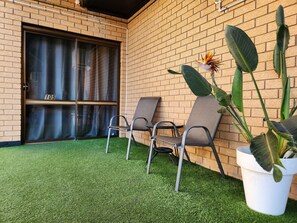 A patio with artificial grass, two metal chairs, a potted plant, and a brick wall.