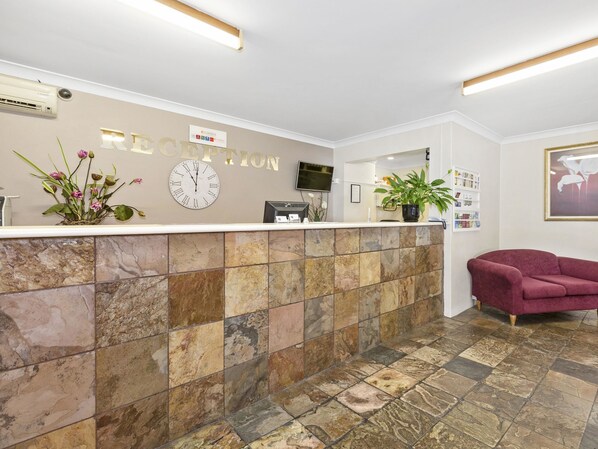 Zona de recepción con un reloj, mostrador de recepción, planta en maceta, sofá y un cuadro en la pared.
