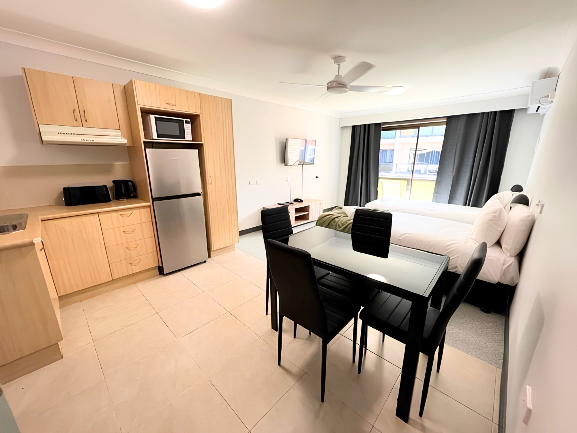 Quarto de hotel moderno com cozinha compacta, área de refeições, cama e uma janela grande com cortinas.
