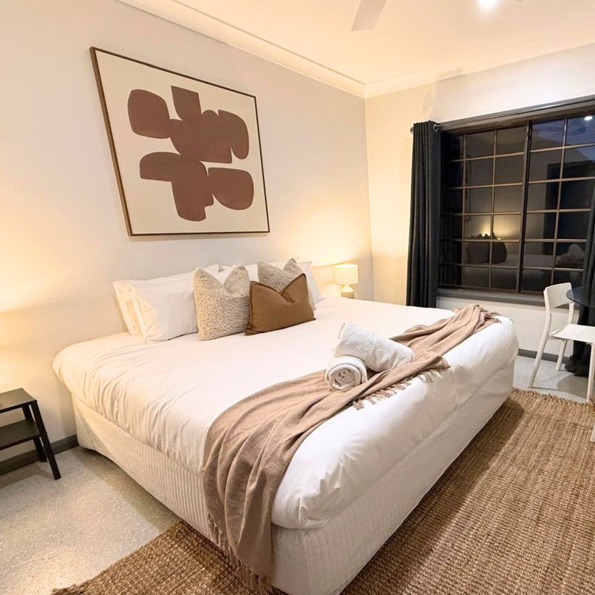 A hotel room with a large bed, a bedside table, a chair, a window with curtains, and a wall-mounted artwork.