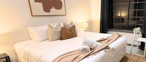 A hotel room with a large bed, a bedside table, a chair, a window with curtains, and a wall-mounted artwork.